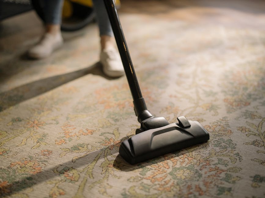 A close-up image of a person using a black vacuum cleaner on a traditional patterned rug with floral motifs in shades of beige, green, and orange, in a well-lit room during daytime. The vacuum cleaner
