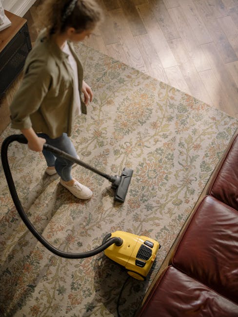 A close-up image of a person using a black vacuum cleaner on a traditional patterned rug with floral motifs in shades of beige, green, and orange, in a well-lit room during daytime. The vacuum cleaner
